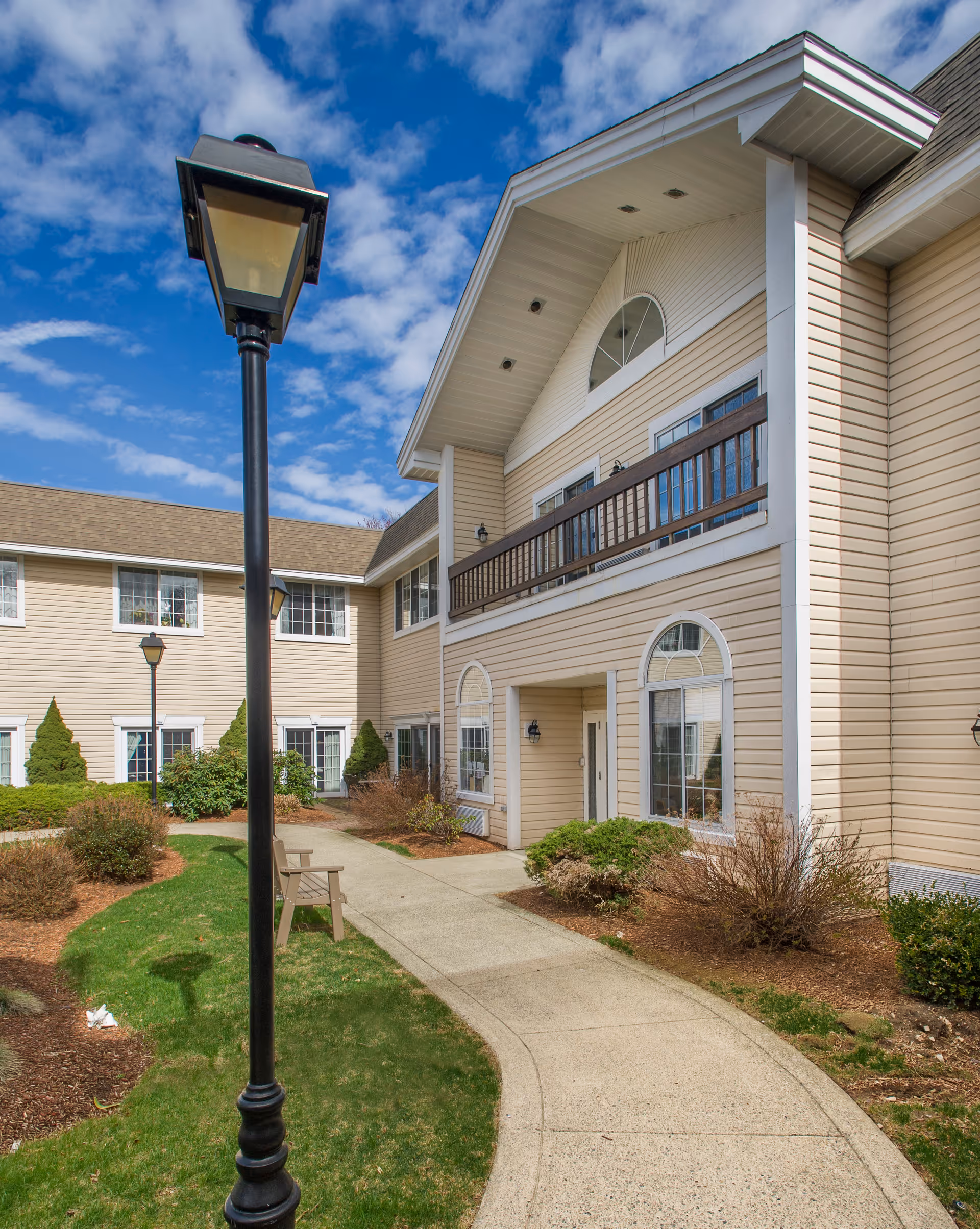Exterior view of a beige two-story senior living facility building with white trim, featuring a balcony, multiple windows, a curved concrete walkway, green grass, bushes, and black lamp posts under a partly cloudy blue sky.