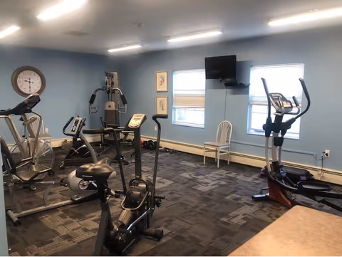 A small fitness room with various exercise equipment including stationary bikes, an elliptical machine, and a multi-function weight machine. The room has light blue walls, two windows with blinds, a wall-mounted TV, a clock, and a single white chair. The floor is carpeted with a patterned design.