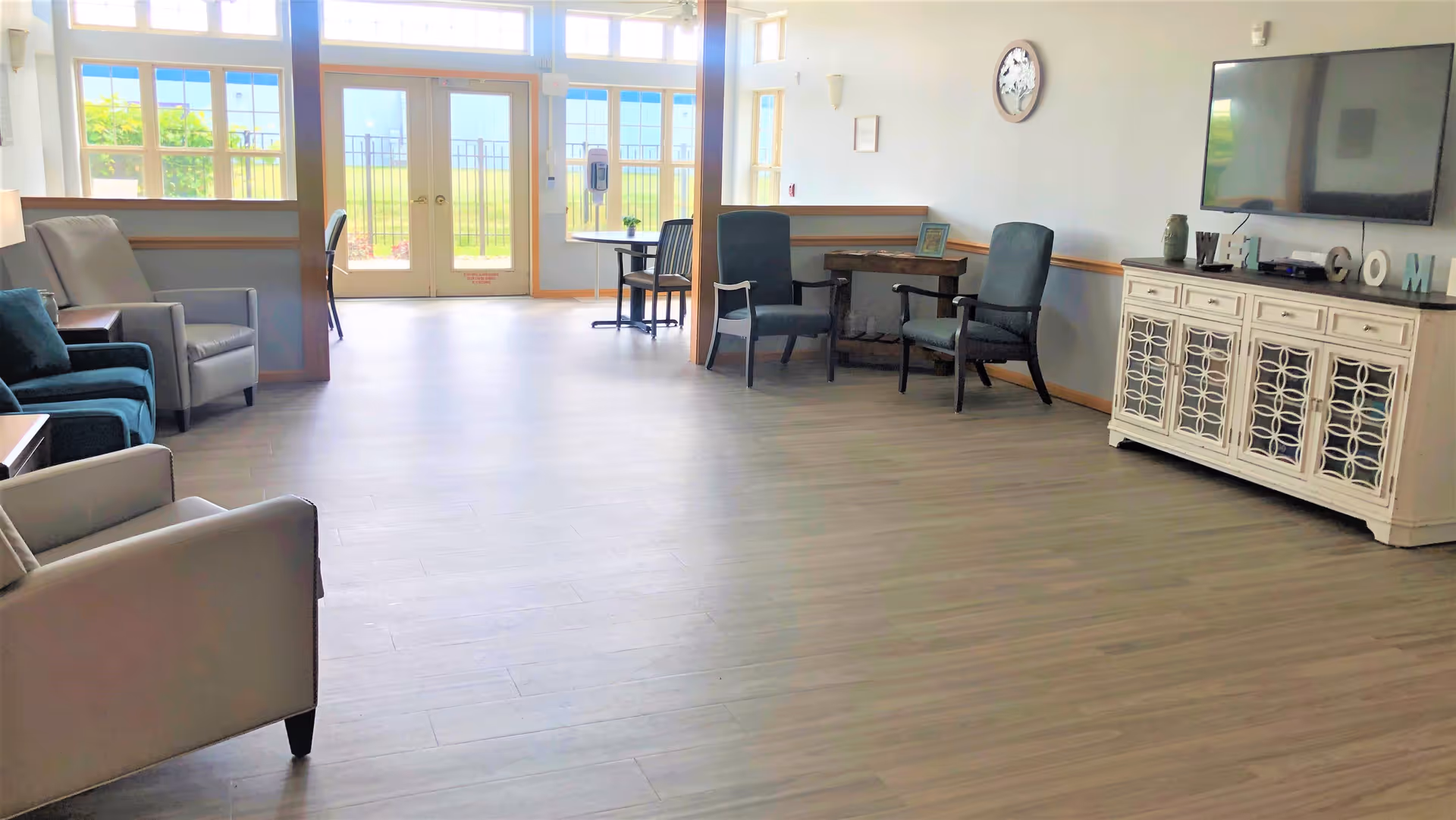 A bright and spacious living room area with multiple armchairs arranged along the walls, a large flat-screen TV mounted on the wall above a white cabinet, and a small table with two chairs near glass double doors leading outside. The room has light-colored wooden flooring and large windows letting in natural light.