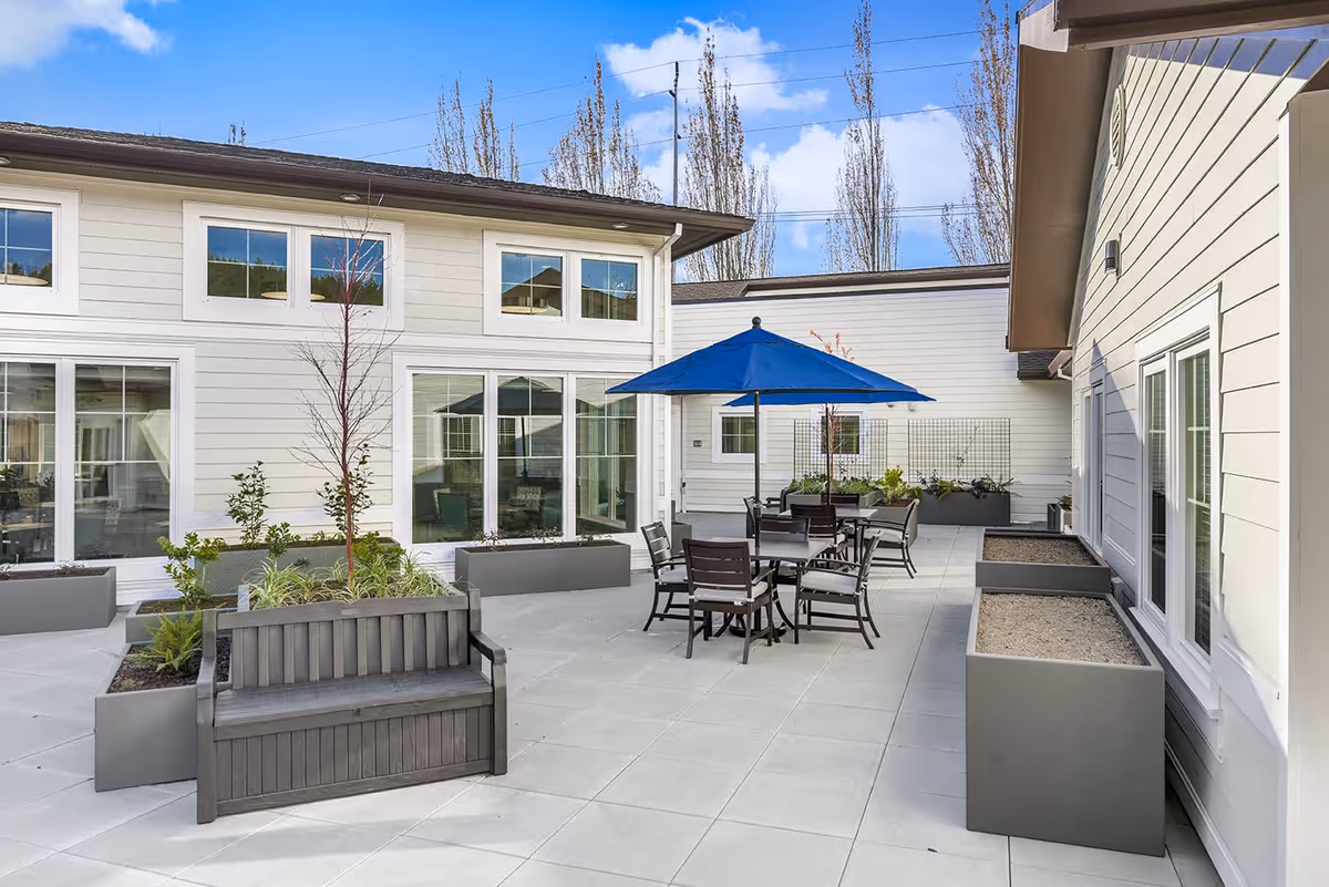 Outdoor patio area at Courtyard at Mt. Tabor with gray tiled flooring, several planter boxes with small plants, a wooden bench, and a table with chairs under a blue umbrella. The patio is surrounded by light-colored building walls with multiple windows and a clear blue sky above.