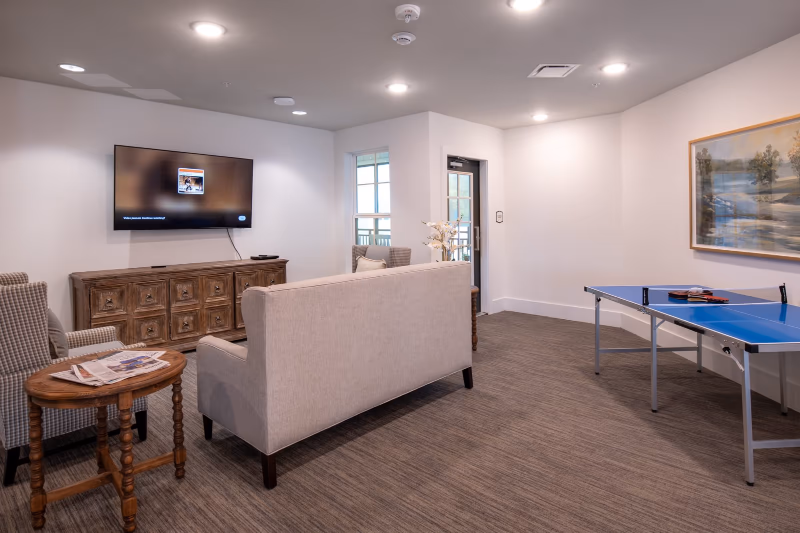 A bright common lounge with a sofa and chairs facing a wall-mounted TV and a ping-pong table to the side.
