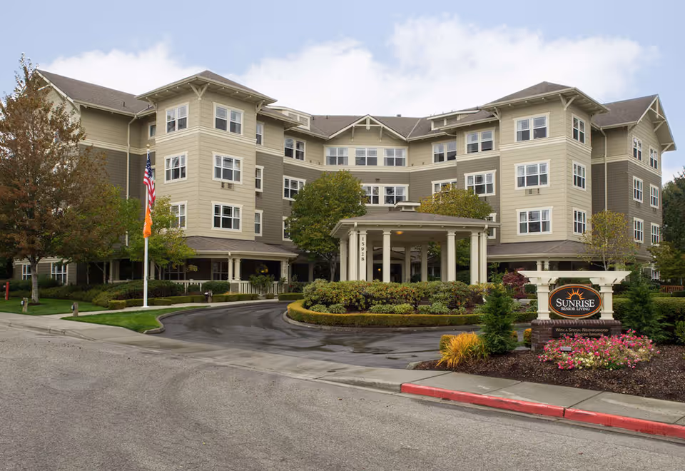 Exterior view of Sunrise of Bellevue senior living facility showing a multi-story building with beige and brown siding, surrounded by landscaped greenery and trees. There is a driveway leading to a covered entrance with columns, and a sign in front displaying the facility's name.