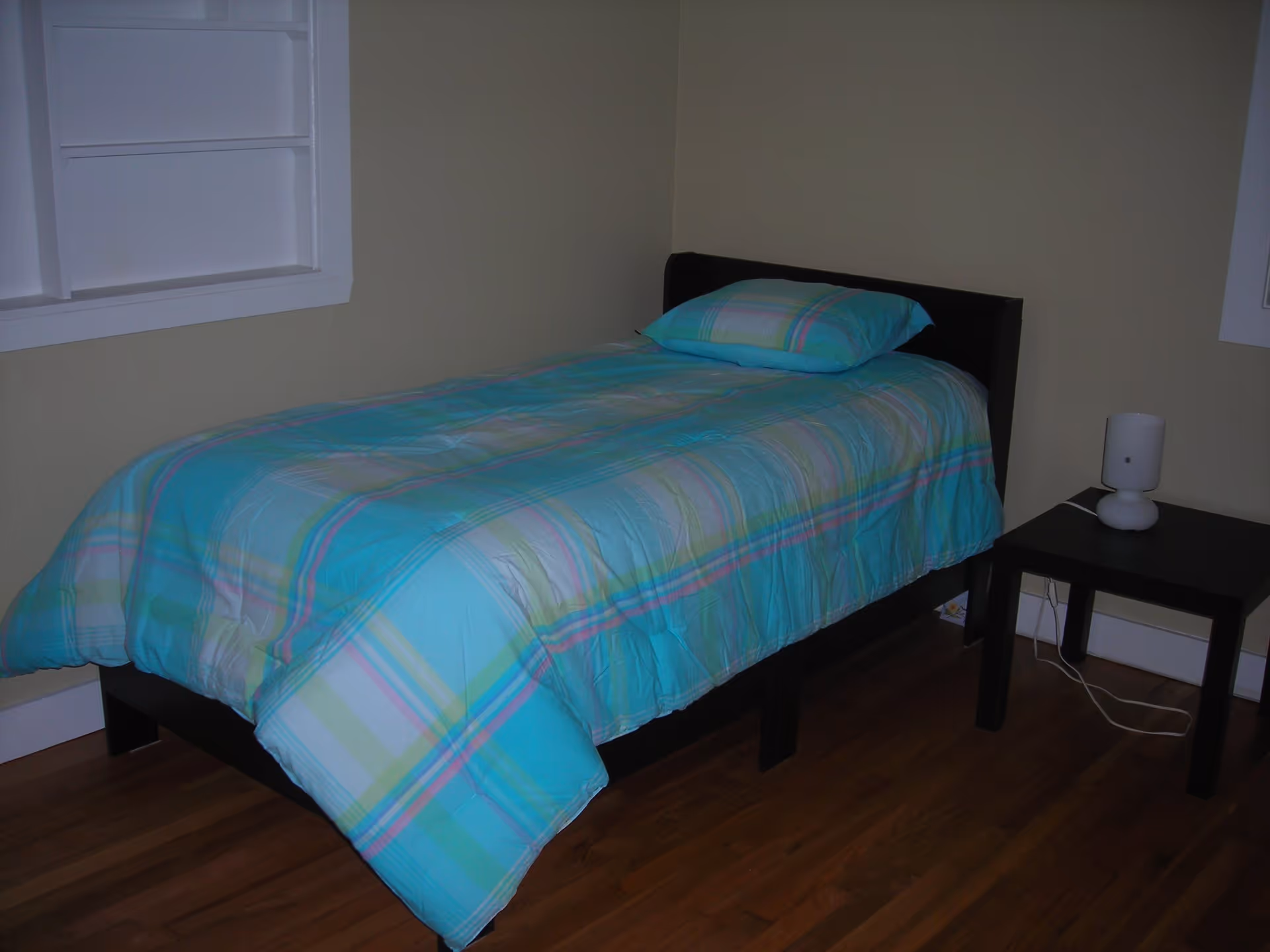 A small bedroom with a single bed covered in a teal plaid comforter and a black nightstand with a lamp.
