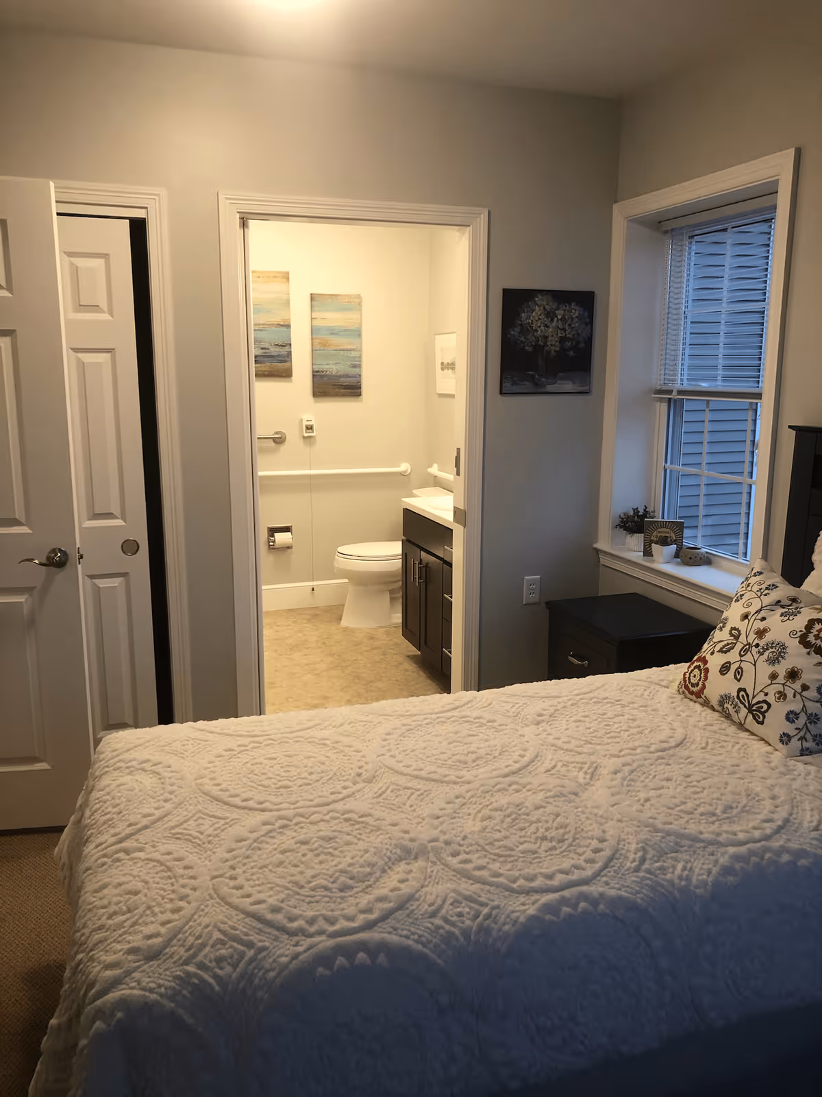 A cozy bedroom with a white textured bedspread and a decorative pillow with floral patterns. To the right is a window with blinds and a small nightstand with decorative items. The bedroom opens into a bathroom with a toilet, sink, and two landscape paintings on the wall. The walls are painted light gray and the room has a calm, inviting atmosphere.