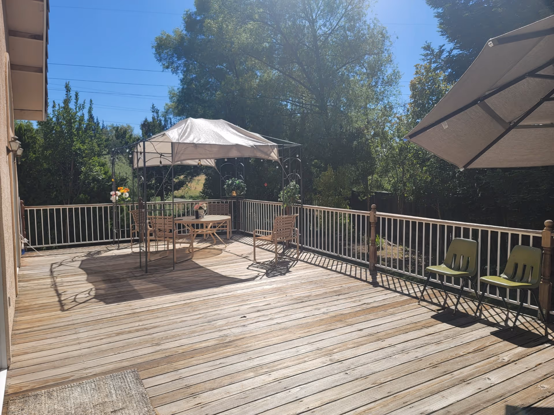 A spacious wooden deck outdoors with a white canopy tent covering a round table and chairs. There are two green chairs under a large umbrella on the right side. The deck is surrounded by a white railing and trees are visible in the background under a clear blue sky.