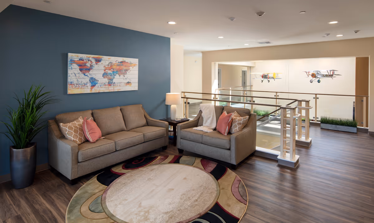 A cozy sitting area with a beige three-seater sofa and a matching loveseat, both adorned with decorative pillows and a throw blanket. A round patterned rug lies on the wooden floor, and a tall green plant in a metallic pot is placed beside the sofa. A side table with a lamp sits between the two sofas. The wall behind the sofa is painted blue and features a colorful world map artwork. The area overlooks a hallway with glass railings and two model airplanes hanging from the ceiling.