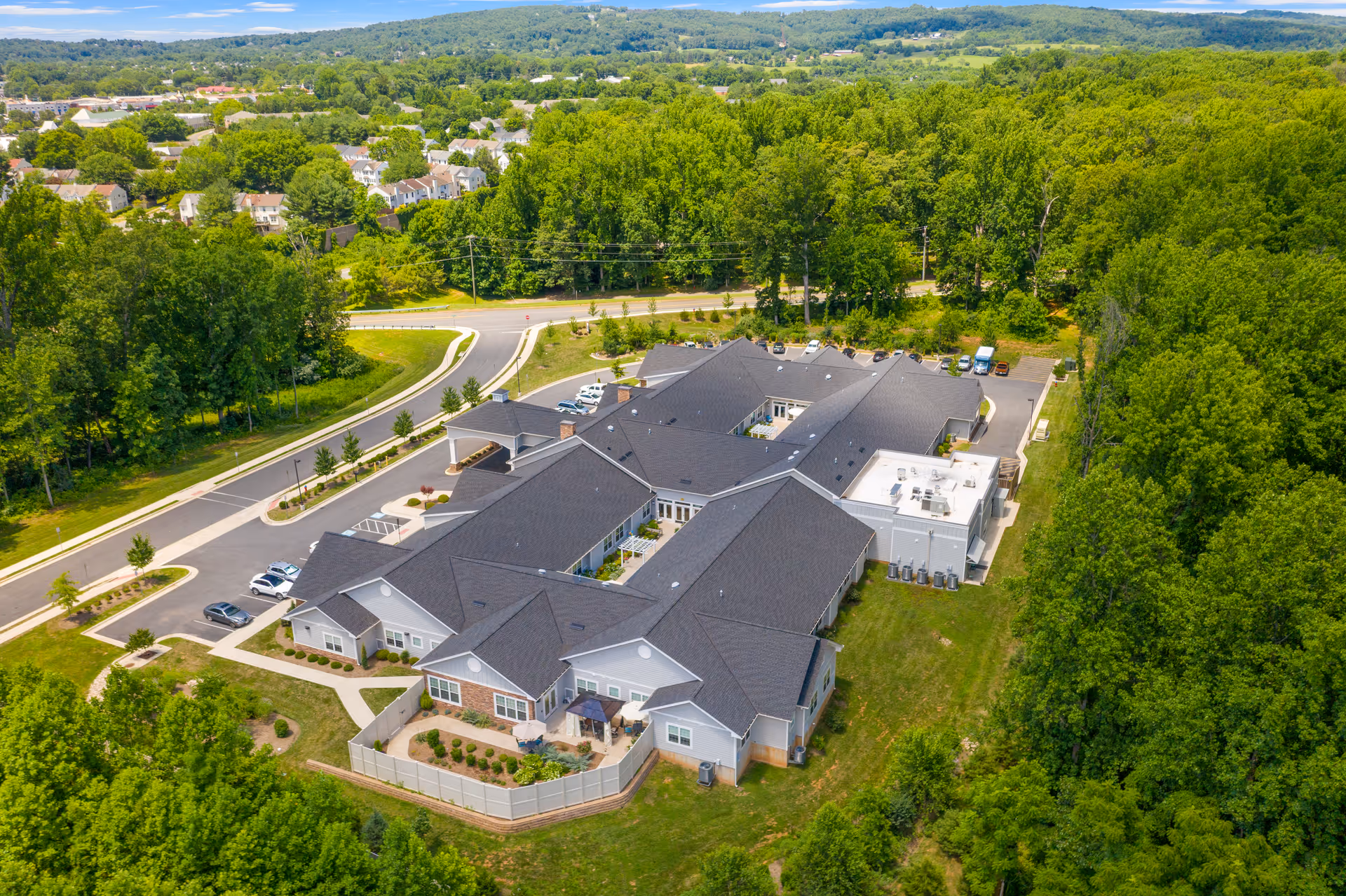 Aerial view of Poet's Walk Warrenton, a memory care community building surrounded by trees and greenery, with a parking lot and roads nearby under a clear blue sky.