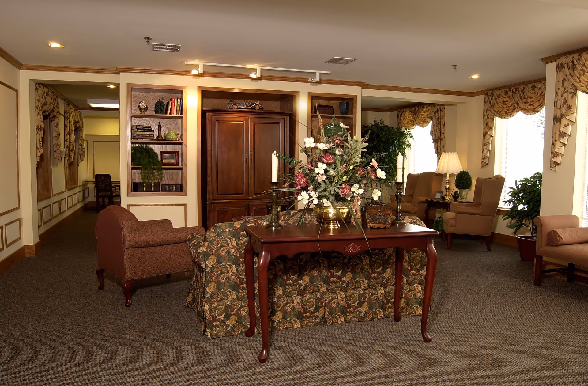 A cozy living room area with floral patterned sofa, a wooden table with a large floral arrangement and candlesticks, armchairs near windows with floral curtains, a wooden cabinet with shelves holding decorative items, and several potted plants. The room has warm lighting and carpeted floor.