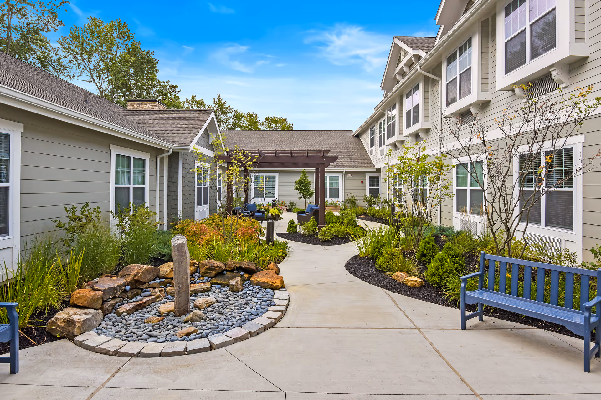 Outdoor courtyard area at The Commons on Meridian featuring a paved walkway, landscaped garden beds with various plants and shrubs, a small rock water feature, blue benches, and a pergola with seating under a clear blue sky.