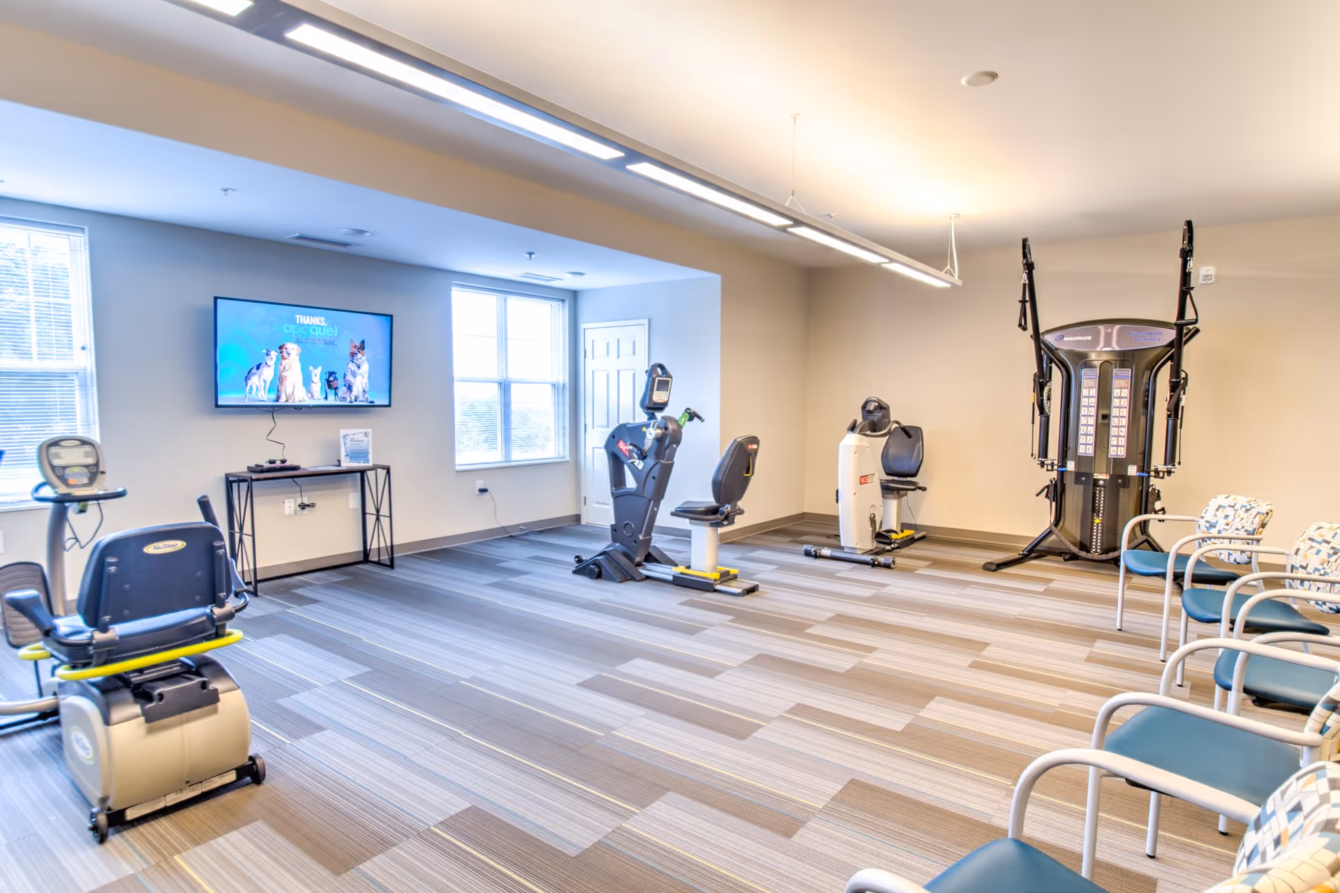 Bright exercise room with recumbent bikes, a multi-station strength machine, a wall-mounted TV and a row of chairs.