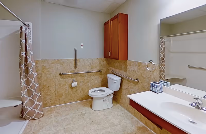 A clean and accessible bathroom with beige tiled walls and floor. It features a white toilet with grab bars on the walls around it, a shower area with a patterned beige and white curtain, a white sink with a countertop, a large mirror above the sink, and a wooden cabinet mounted on the wall.