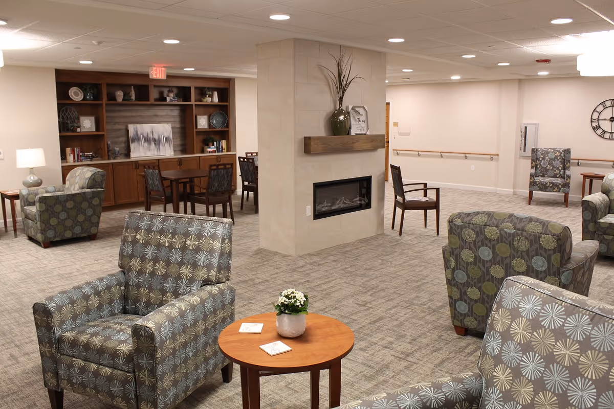 A cozy and well-lit common area in a senior living facility featuring patterned armchairs arranged around small wooden tables. A central column with a built-in fireplace and decorative vase divides the space. In the background, there is a wooden bookshelf with decorative items and a dining table with chairs. The room has carpeted floors and neutral-colored walls with recessed lighting.