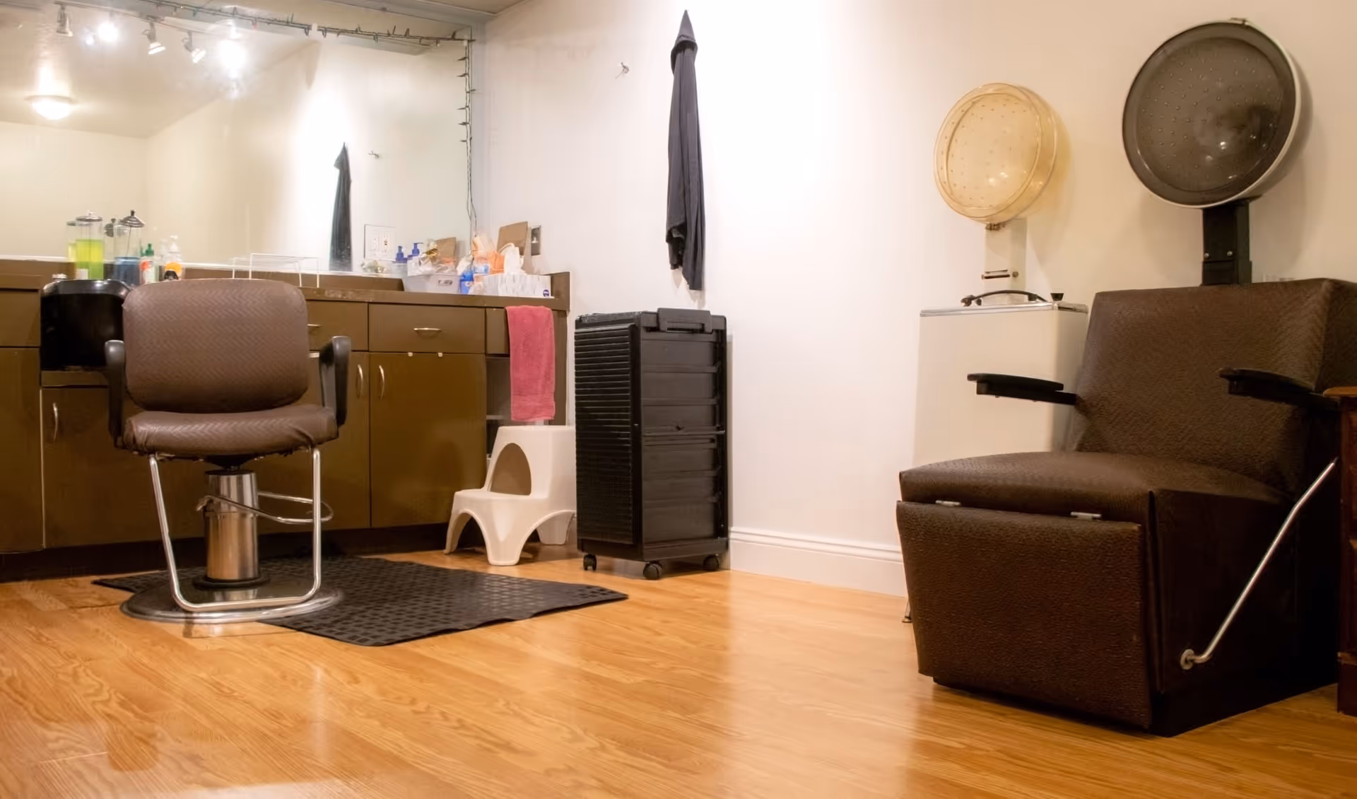 Interior view of a salon area with a brown salon chair on a black mat in front of a counter with cabinets and a large mirror. On the right side, there is a brown hair dryer chair with a hooded hair dryer above it. The floor is wooden and the walls are white.