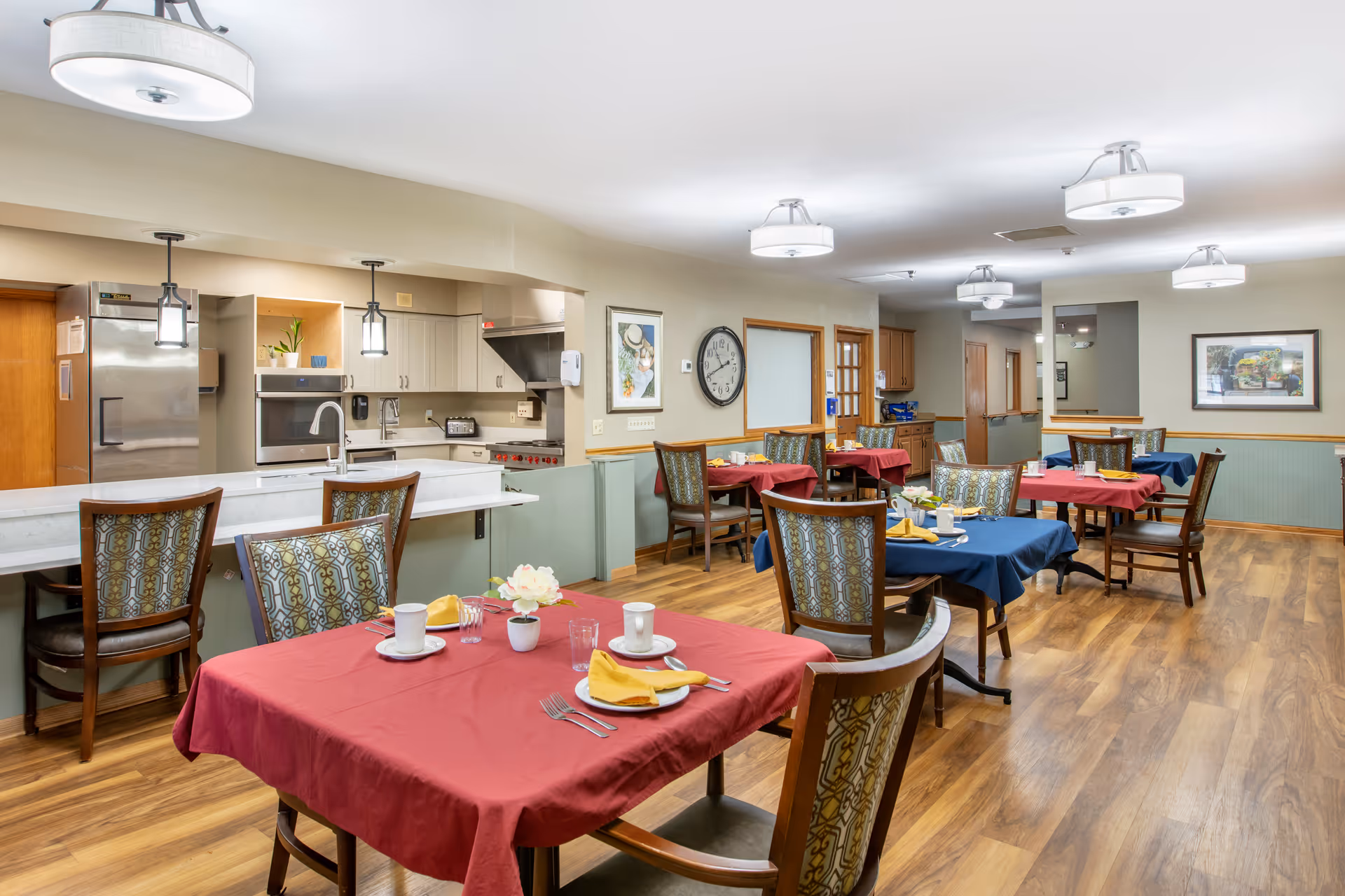 Dining area in Brookdale Saginaw with several tables covered in red and blue tablecloths, each set with plates, cups, utensils, and yellow napkins. The room has wooden flooring, patterned chairs, and a kitchen area with stainless steel appliances and pendant lights in the background.