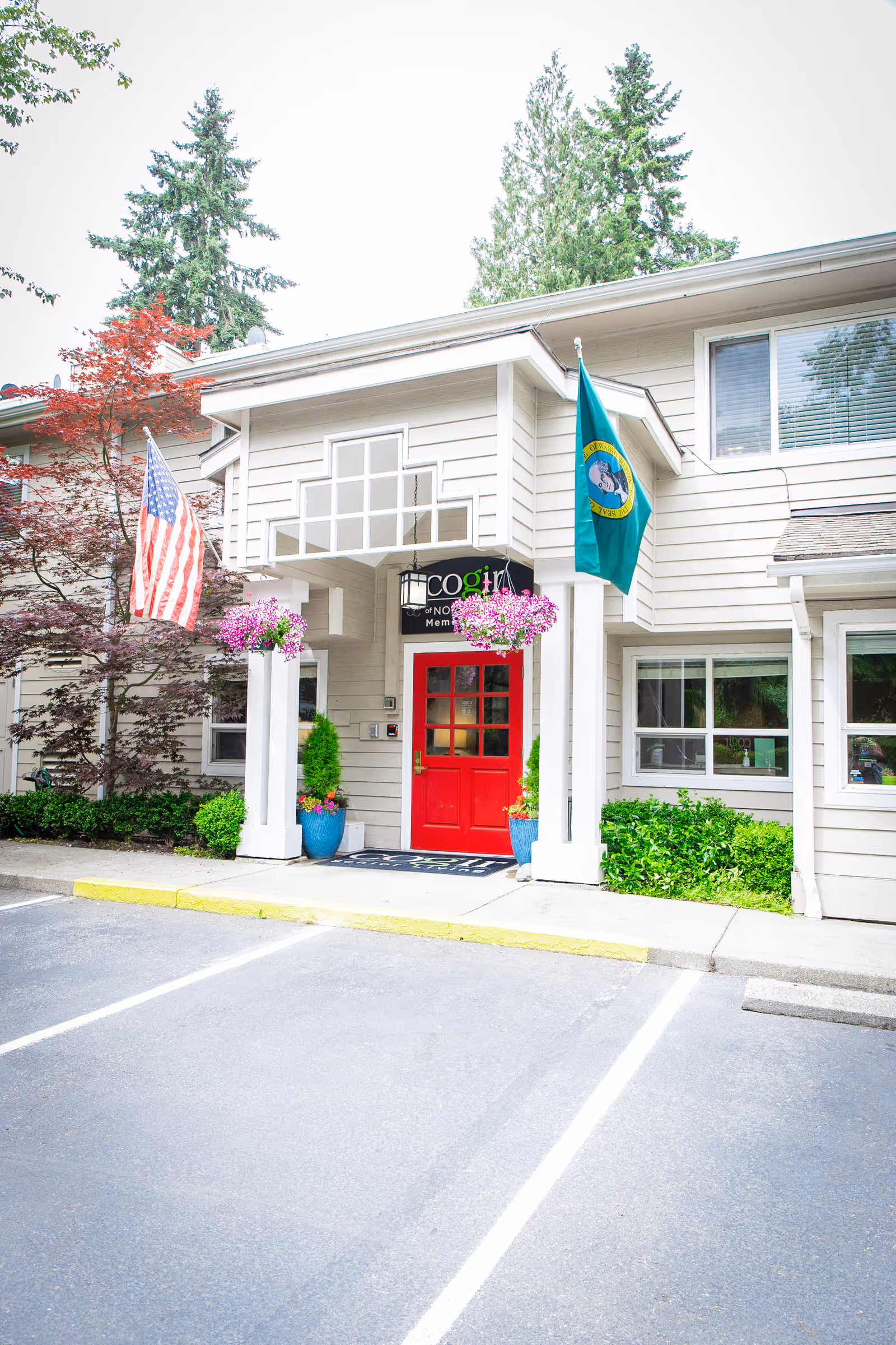 Entrance of Cogir of Northgate facility featuring a red door under a white porch with hanging flower baskets, American and Washington state flags, and surrounding greenery.