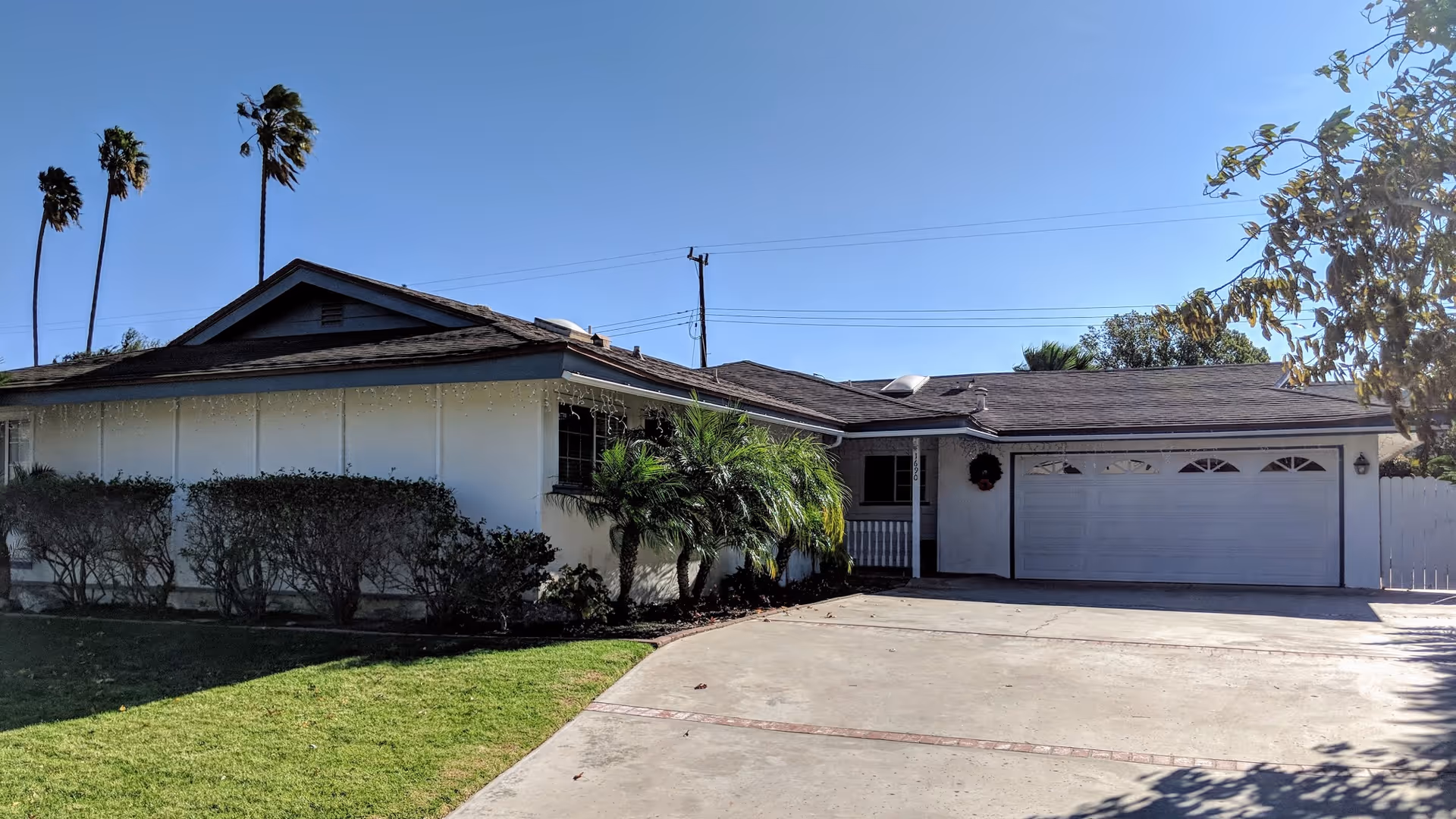 Single-story residential building with a two-car garage, white exterior walls, and a sloped roof. The front yard has trimmed bushes, a small palm tree, and a green lawn. Tall palm trees and a clear blue sky are visible in the background.