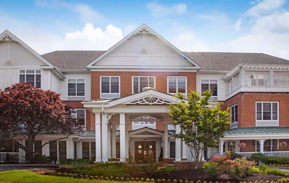 Front entrance of a three-story red brick and white senior living building with columns, a covered porte-cochère, and landscaped grounds.