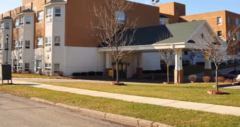 Exterior view of a multi-story assisted living facility building with a covered entrance, leafless trees, and a sidewalk in front.