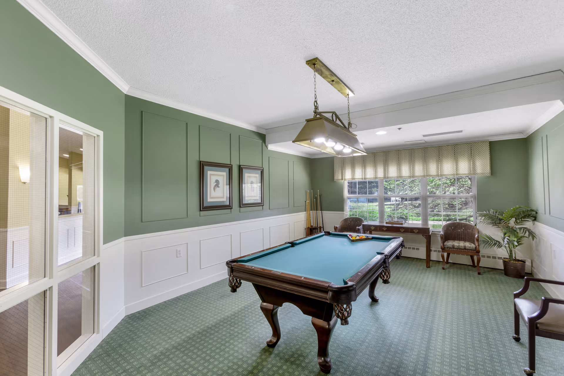 A well-lit game room with a green pool table in the center, surrounded by green walls with white wainscoting. There are two framed pictures on the wall, a window with patterned valance letting in natural light, two wicker chairs, a small table, and a potted plant. A hanging light fixture is above the pool table.