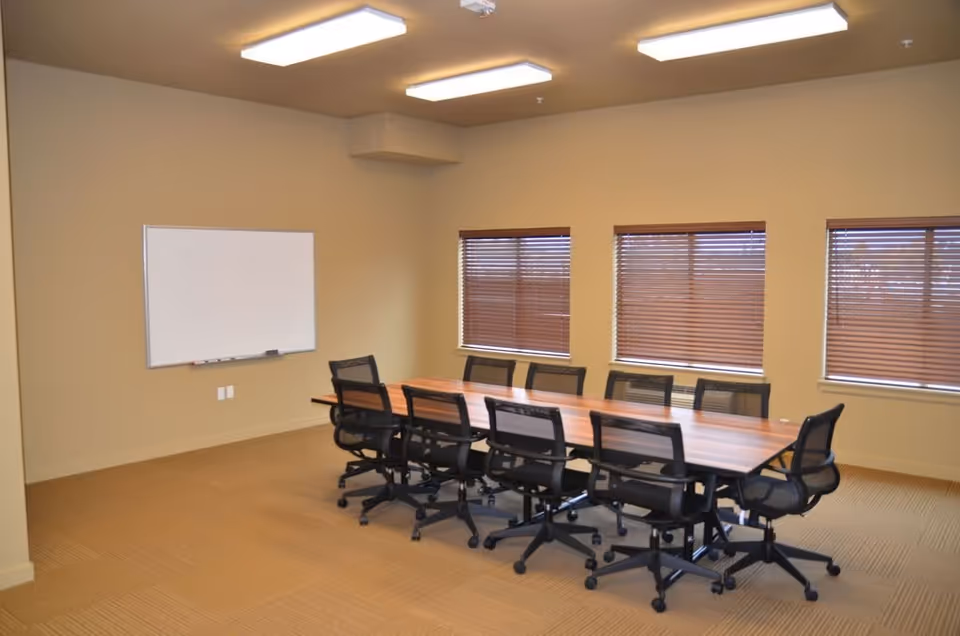 A conference room with a long wooden table surrounded by ten black office chairs on wheels. The room has beige walls, three windows with closed brown blinds, a whiteboard mounted on one wall, and ceiling lights.