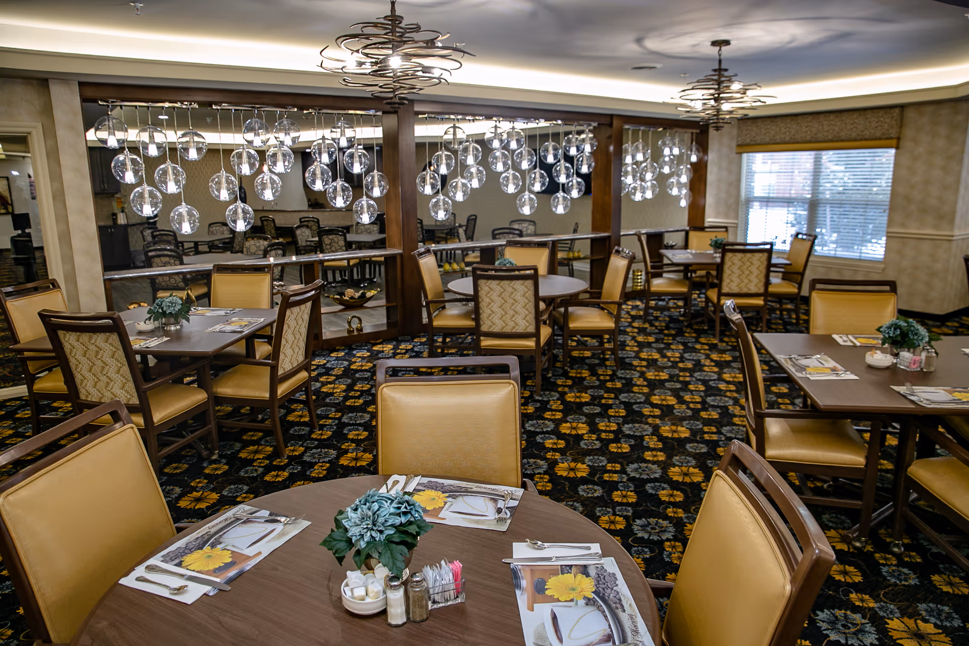 Dining room with multiple round and square tables set with placemats, utensils, and small flower centerpieces. The room features patterned carpet, large mirrors on one wall, modern hanging light fixtures with glass globes, and windows with blinds allowing natural light.