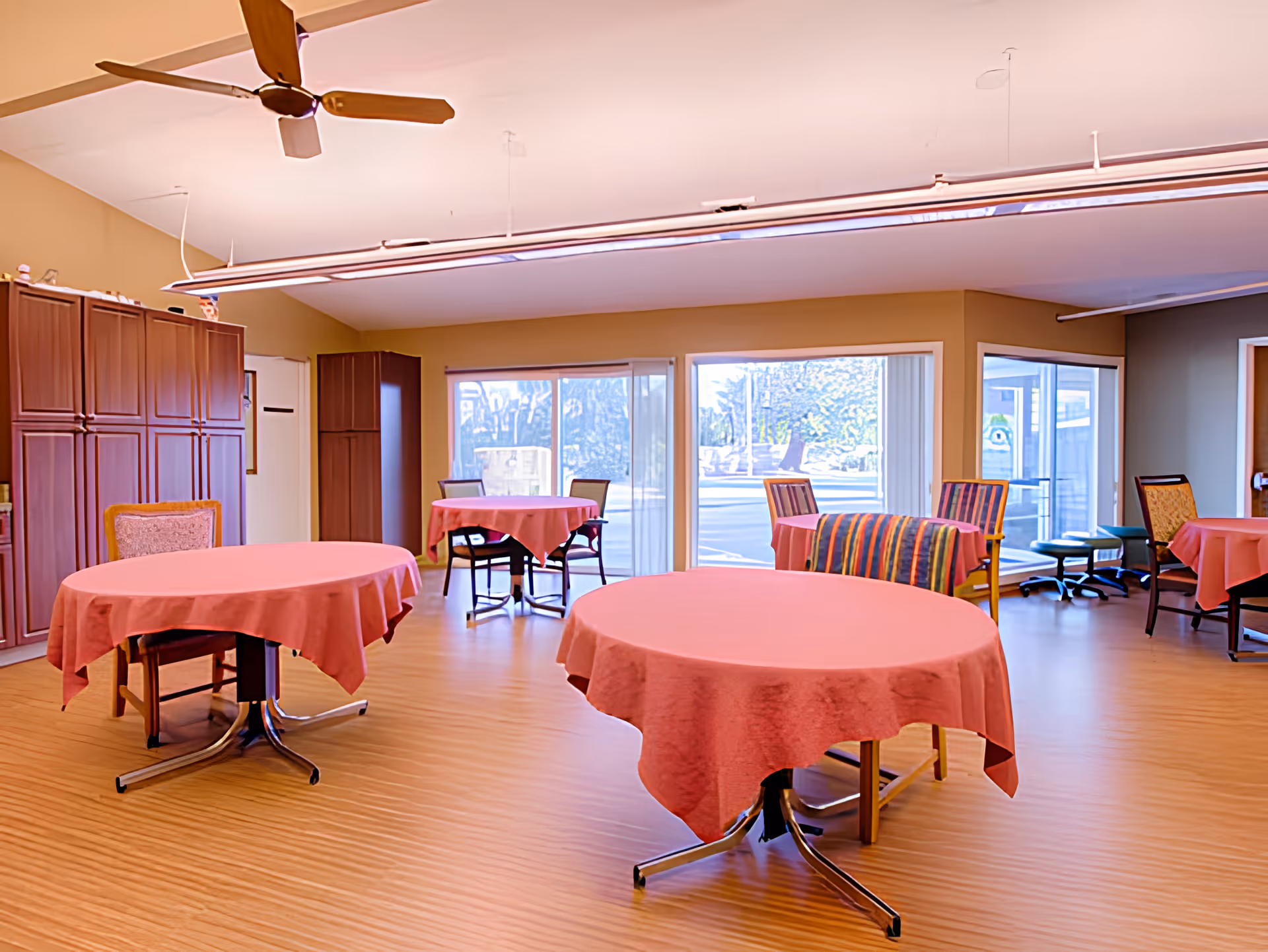 A spacious dining room with round tables covered in pink tablecloths and chairs arranged around them. The room has large windows letting in natural light, wooden flooring, and a ceiling fan. There are cabinets along one wall and a few chairs with colorful upholstery near the windows.