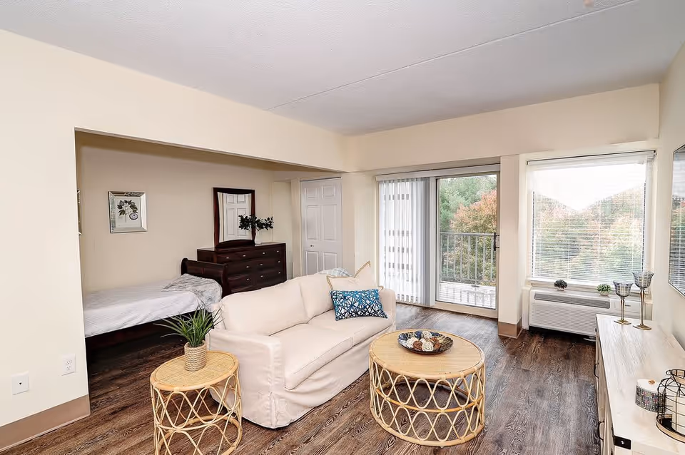 A bright and cozy living space featuring a white sofa with decorative pillows, two round wicker coffee tables, a wooden dresser with a mirror, and a bed partially visible in an alcove. Large windows and a sliding glass door provide natural light and a view of trees outside. The room has wood flooring and light-colored walls.