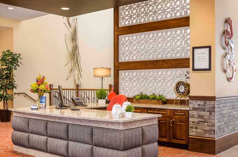 A reception desk in a senior living lobby with decorative wall panels, plants, lamps, and desk accessories.