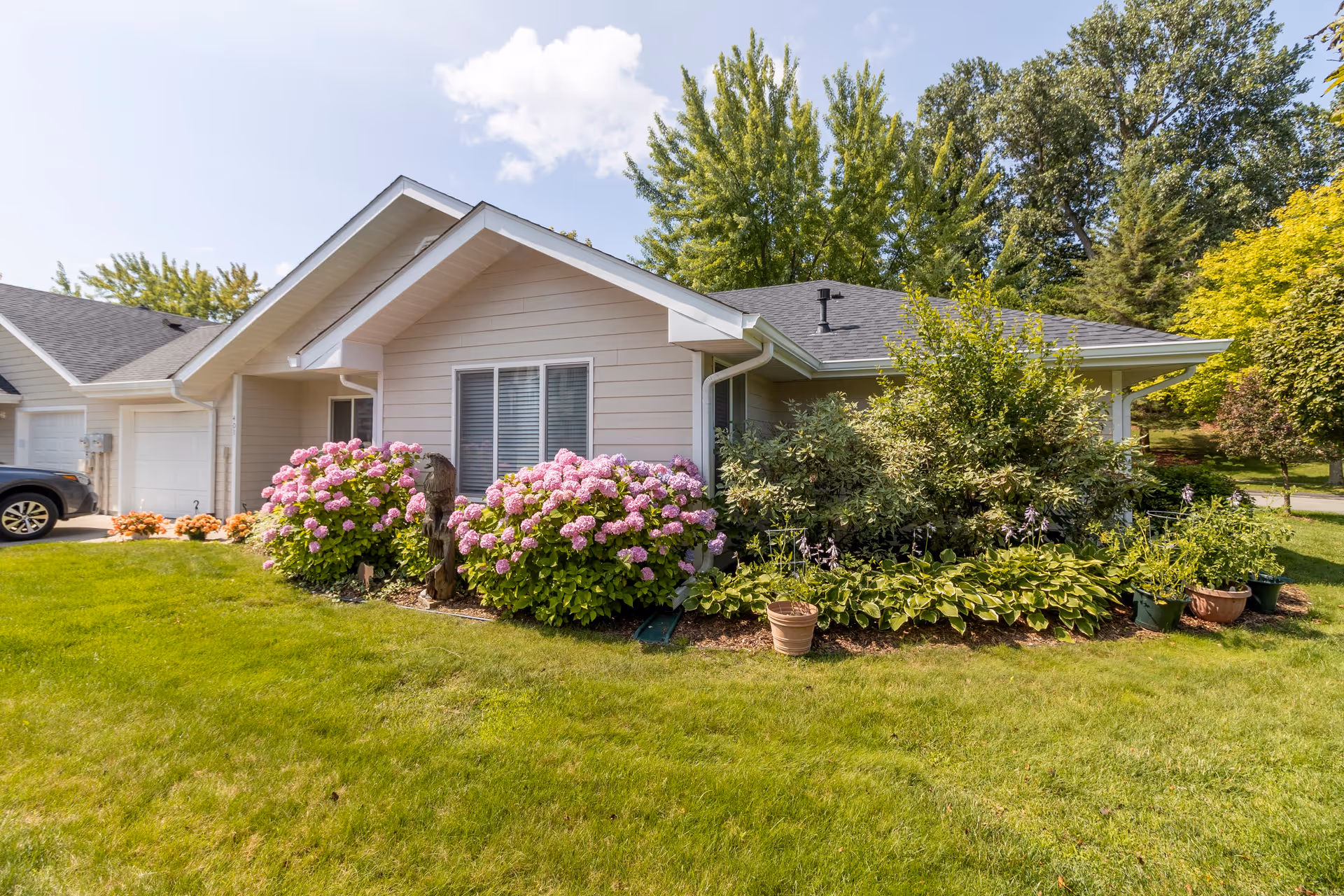 Exterior view of a single-story residential building with beige siding and a gray roof, surrounded by a well-maintained lawn and landscaped garden featuring pink hydrangeas, green shrubs, and potted plants under a partly cloudy sky.