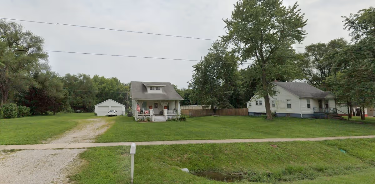 A small white house with a front porch and an American flag on the lawn, situated on a large grassy lot with a gravel driveway leading to a detached garage. Another white house is visible to the right, with trees surrounding the area under a cloudy sky.