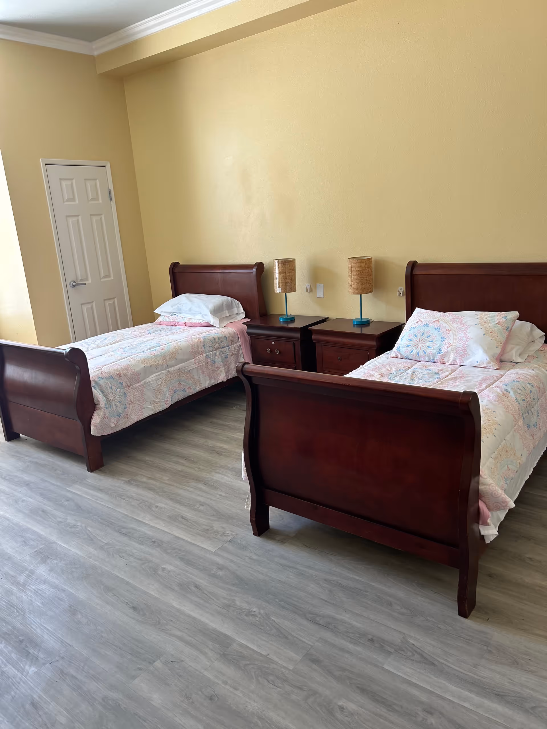Two twin beds with dark wooden sleigh frames, a shared nightstand and lamps in a bright, simply furnished bedroom.