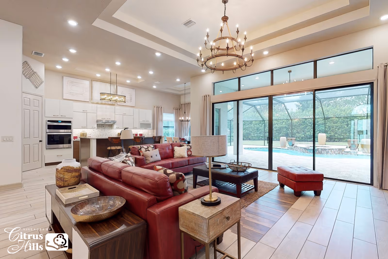 Open-plan living room with red leather sofas, coffee table and chandelier opening via large sliding glass doors to a pool and visible kitchen.