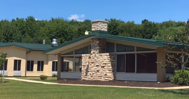 Exterior view of a single-story building with a green roof and a prominent stone chimney, surrounded by a well-maintained lawn and trees in the background under a clear blue sky.