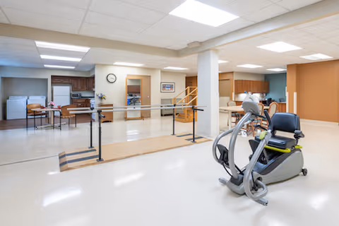 A spacious rehabilitation room with exercise equipment including a stationary recumbent bike and parallel bars for walking practice. The room has a clean, bright interior with white floors, beige walls, and a ceiling with fluorescent lighting. In the background, there is a kitchenette area with cabinets, a refrigerator, and a washing machine and dryer. A clock and framed pictures are mounted on the walls.