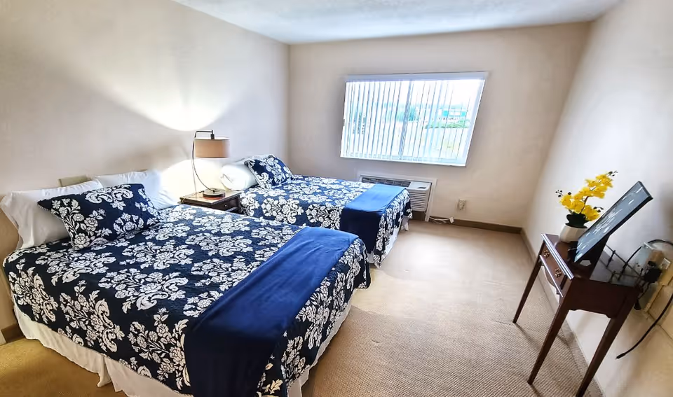 A bedroom with two twin beds covered in matching dark blue and white floral patterned bedspreads and navy blue throws. Between the beds is a small wooden nightstand with a lamp. A window with vertical blinds lets in natural light. On the right side of the room, there is a small wooden table with a vase of yellow flowers and a framed picture. The room has beige walls and carpeted floor.