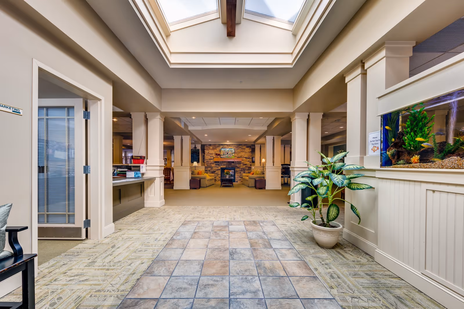 Interior view of a senior living facility lobby area with a tiled floor leading to a carpeted seating area with sofas and a stone fireplace. There is a large skylight overhead, a potted plant on the right, and a large aquarium built into the wall. A door labeled 'Marketing' is visible on the left.