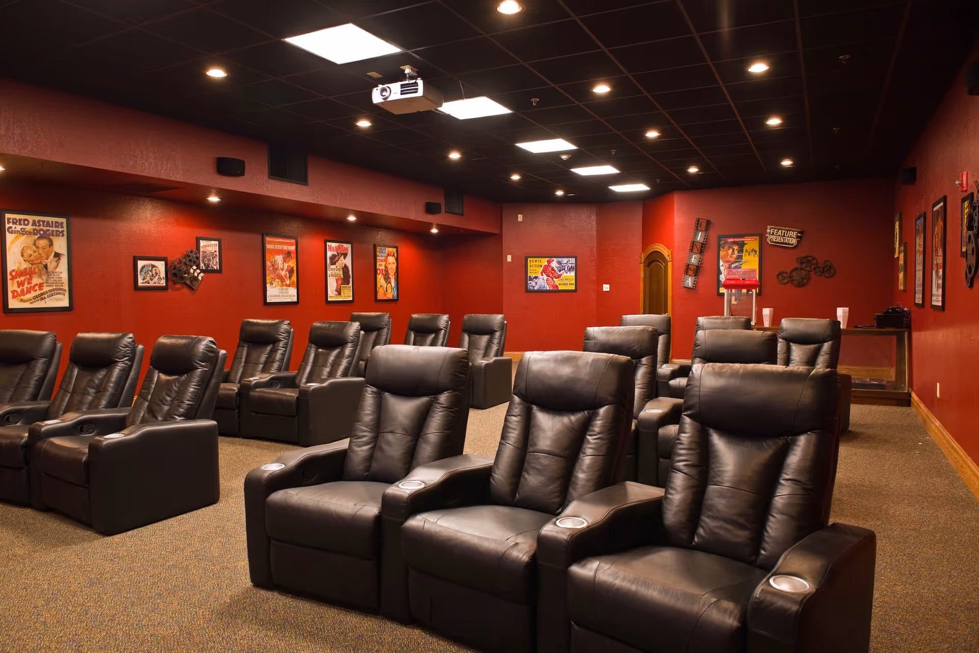 A cozy movie theater room with multiple rows of black leather recliner chairs. The walls are painted dark red and decorated with vintage movie posters. The ceiling has recessed lighting and a projector is mounted in the center.