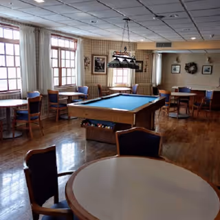 A spacious room with wooden floors featuring a pool table in the center surrounded by multiple round tables and chairs. Large windows with white curtains allow natural light to fill the room. The walls are decorated with framed pictures and a wreath.