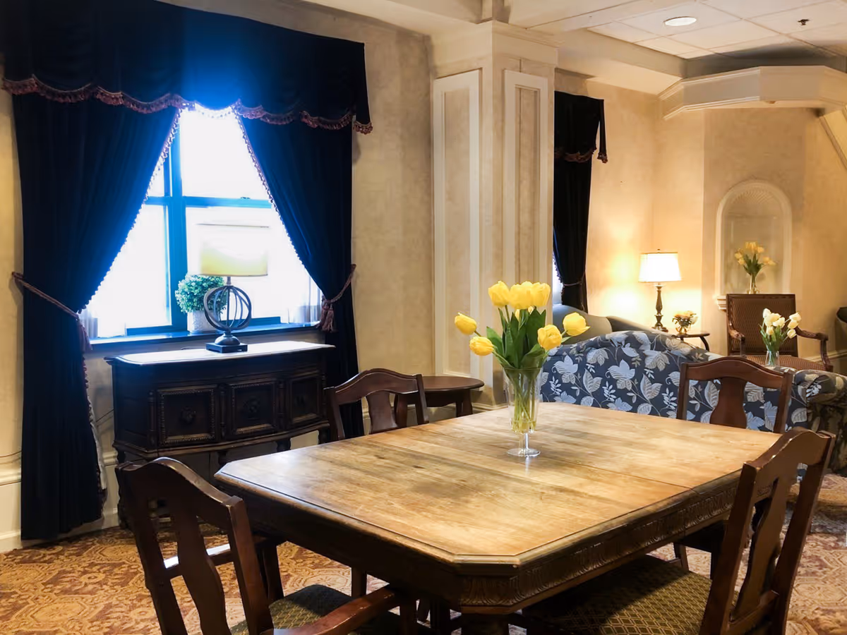 A cozy living room area with a wooden dining table and chairs in the foreground, decorated with a vase of yellow tulips. In the background, there are upholstered chairs and sofas with floral patterns, a side table with a lamp, and a window with dark curtains letting in natural light.