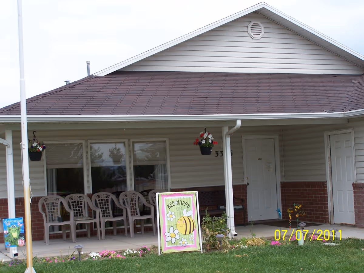 Front porch of a single-story assisted living building with chairs, hanging flower baskets, and a decorative 'Bee Happy' sign on the lawn.