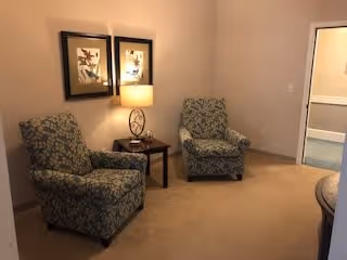 A cozy interior seating area with two patterned armchairs placed around a small wooden side table with a decorative lamp. Two framed floral artworks hang on the wall behind the chairs. The room has beige carpeting and light-colored walls, with a doorway visible on the right side.