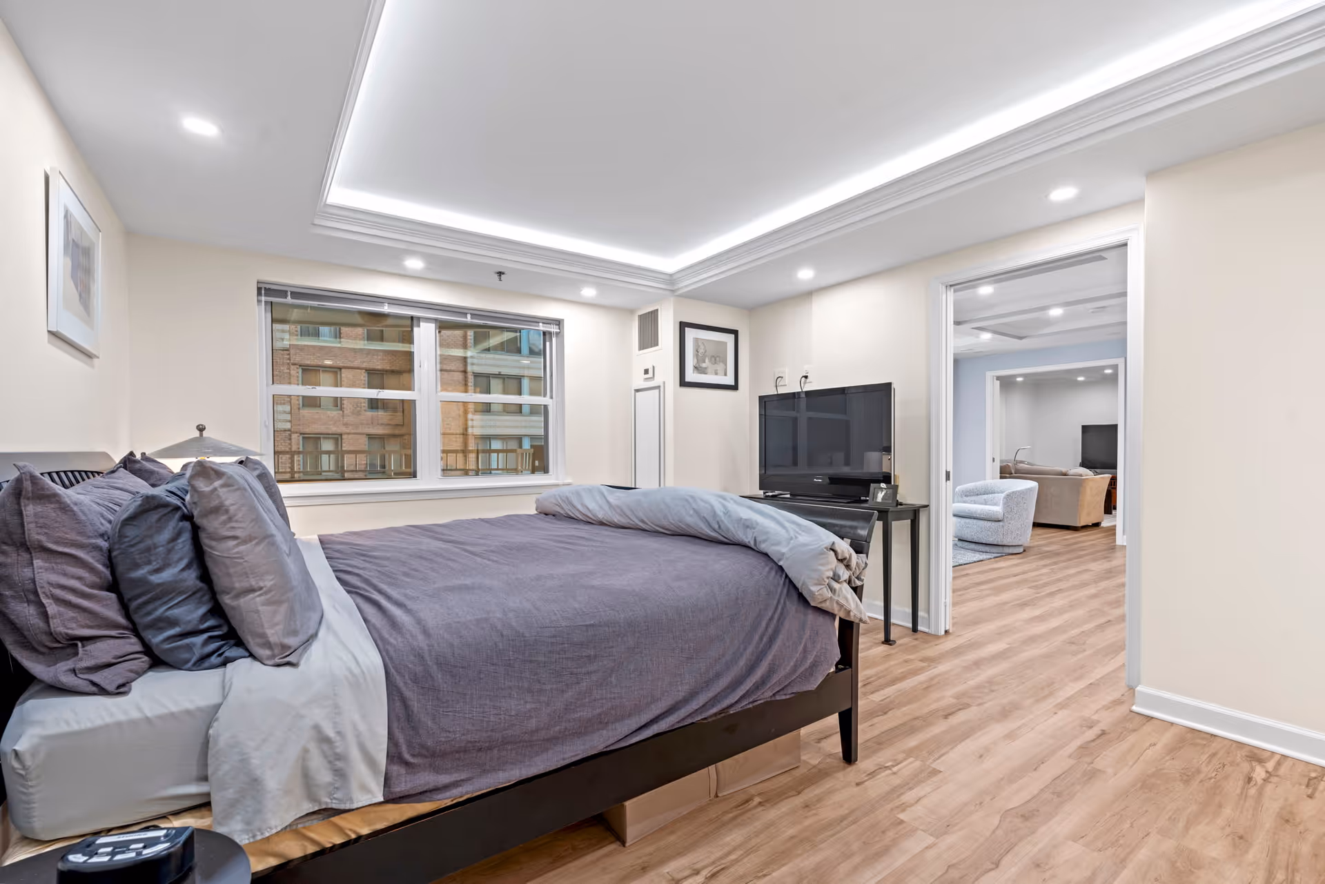 A bedroom with a neatly made bed featuring gray and purple bedding. The room has light-colored walls and wood flooring. A window shows a view of a brick building outside. A flat-screen TV is placed on a small black table against the wall. Through an open doorway, a living room area with chairs and a sofa is visible.