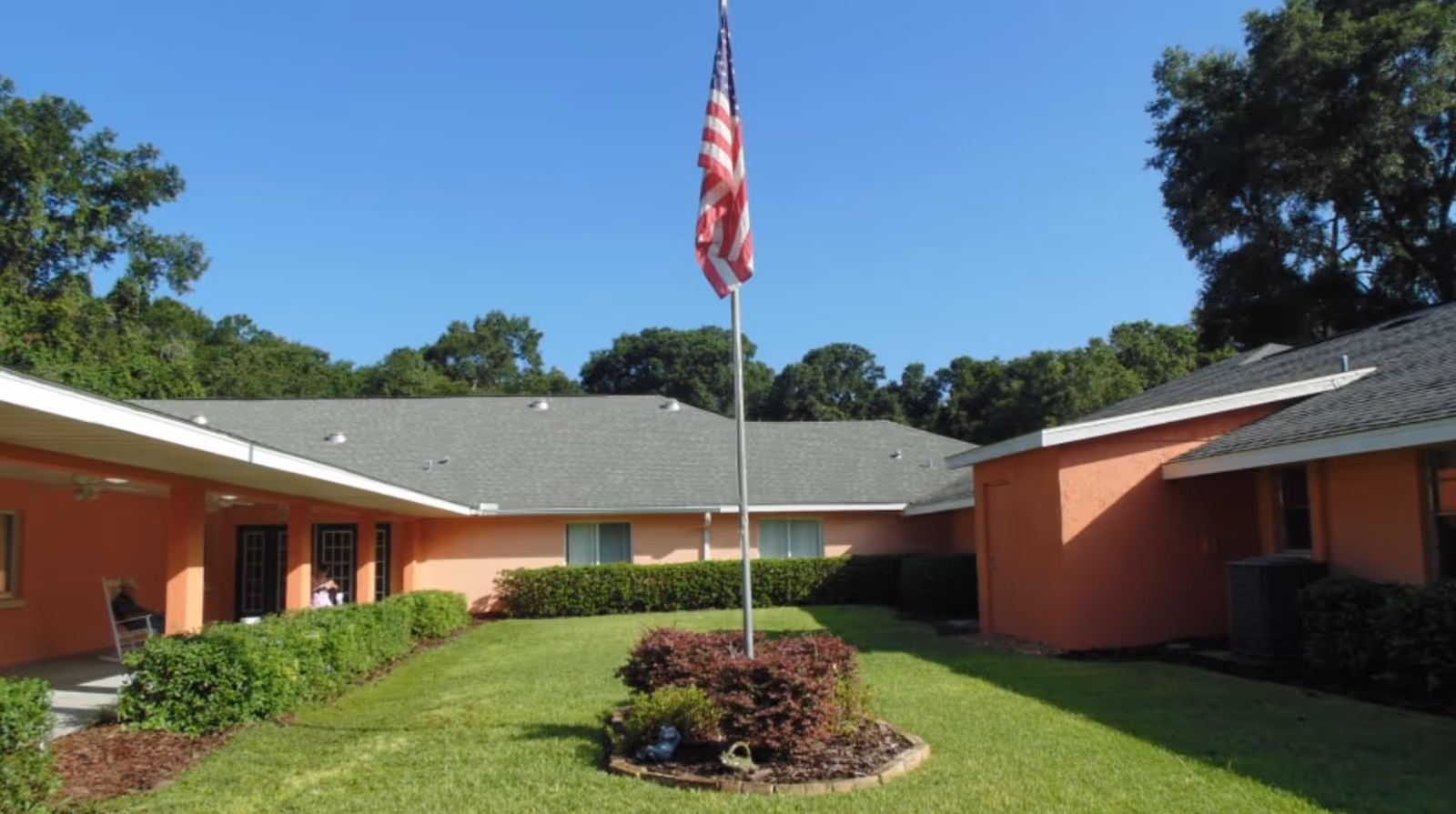 Outdoor courtyard area of a senior living facility with a green lawn, neatly trimmed bushes, and an American flag on a flagpole in the center. The building surrounding the courtyard is painted peach with a gray roof, and there are trees in the background under a clear blue sky.