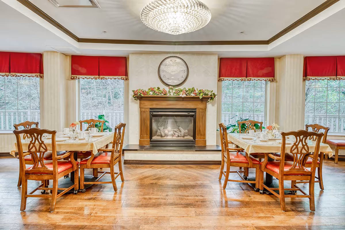 A bright dining room with wooden floors, two tables each surrounded by four wooden chairs with red cushions. The tables are set with white tableware and small flower arrangements. A fireplace with a wooden mantle decorated with greenery and flowers is centered between two large windows with red valances. A large round clock hangs above the fireplace, and a crystal chandelier is mounted on the ceiling.