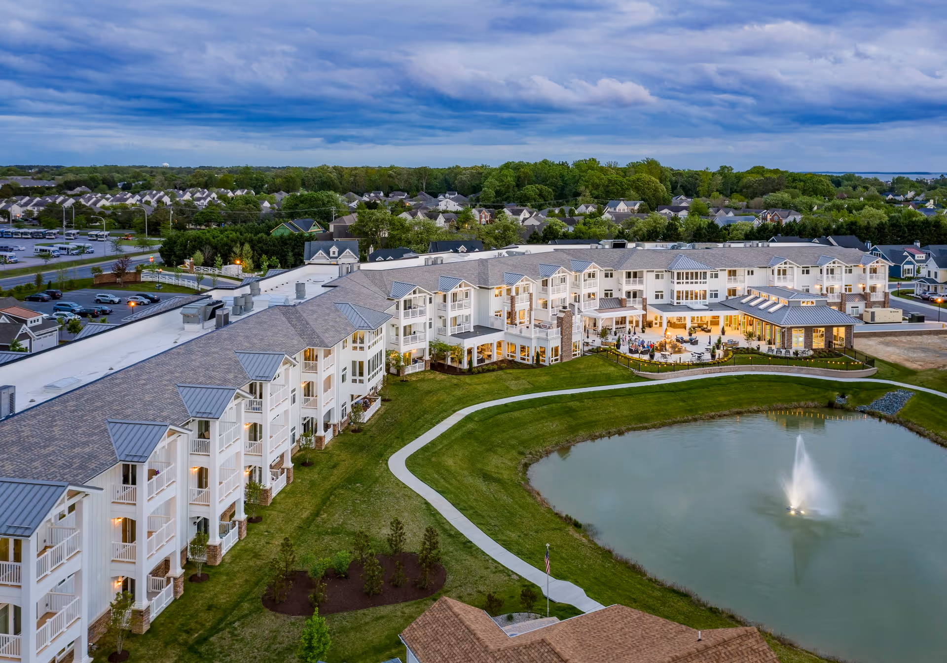 Aerial view of The Lodge at Truitt Homestead senior living facility at dusk, showing a large multi-story building with balconies, a landscaped lawn, a walking path around a pond with a fountain, and outdoor seating areas with people gathered near a fire pit.
