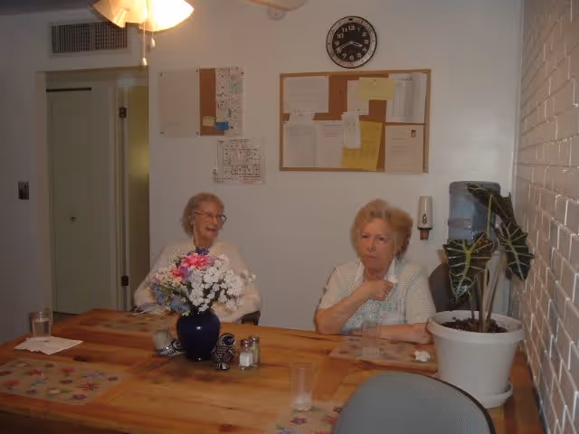 Two elderly women sitting at a wooden table in a room with white walls. On the table is a blue vase with a bouquet of flowers, salt and pepper shakers, and placemats. Behind them on the wall are two bulletin boards with papers pinned and a round clock showing the time as 11:10. There is a large potted plant on the right side of the table and a water dispenser next to it.