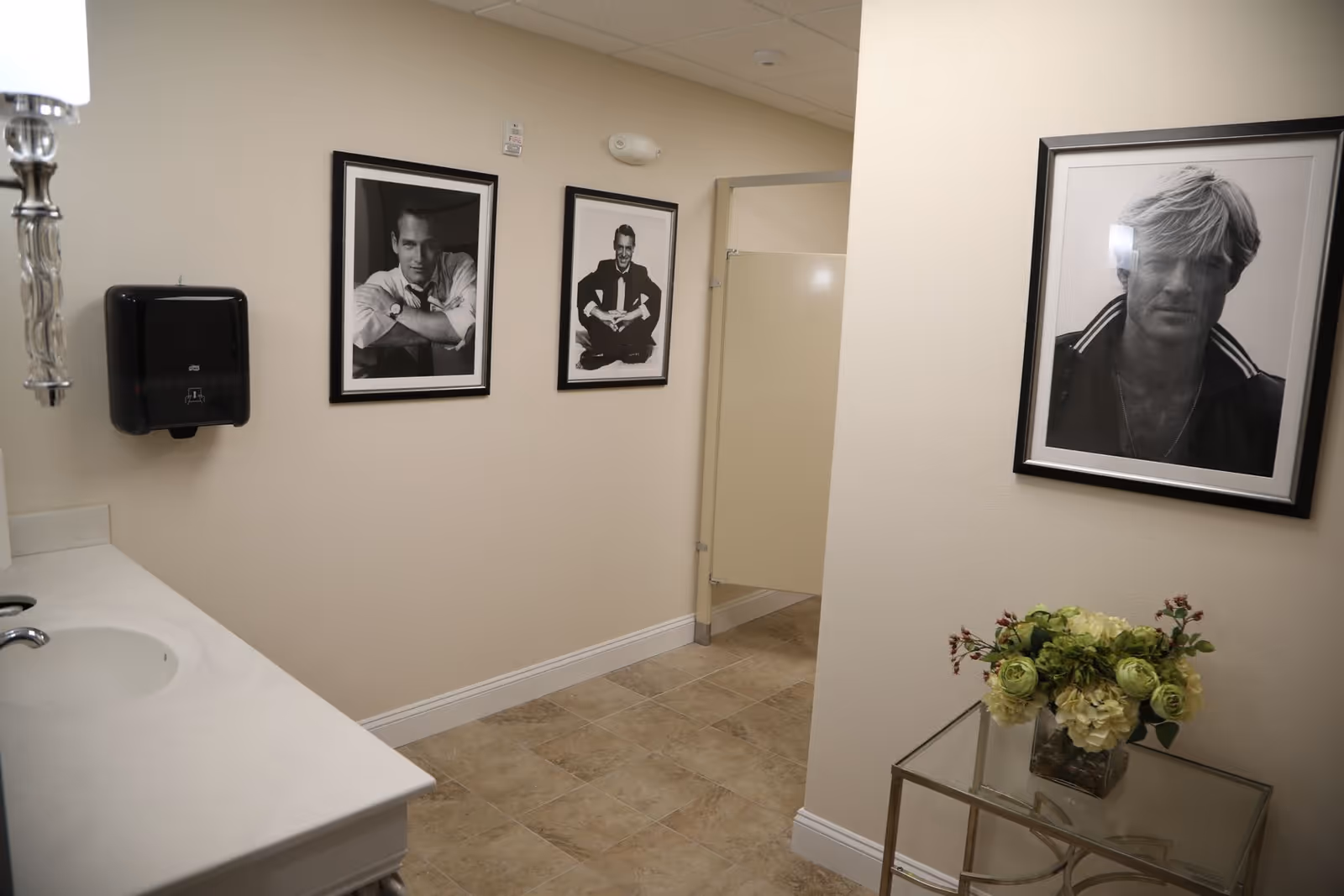 Interior view of a bathroom with beige tiled floor and cream-colored walls. There is a white countertop with a sink on the left side, a black paper towel dispenser mounted on the wall, and three black and white framed photographs of men hanging on the walls. A beige bathroom stall is visible in the background. On the right side, there is a glass table with a floral arrangement in a vase.