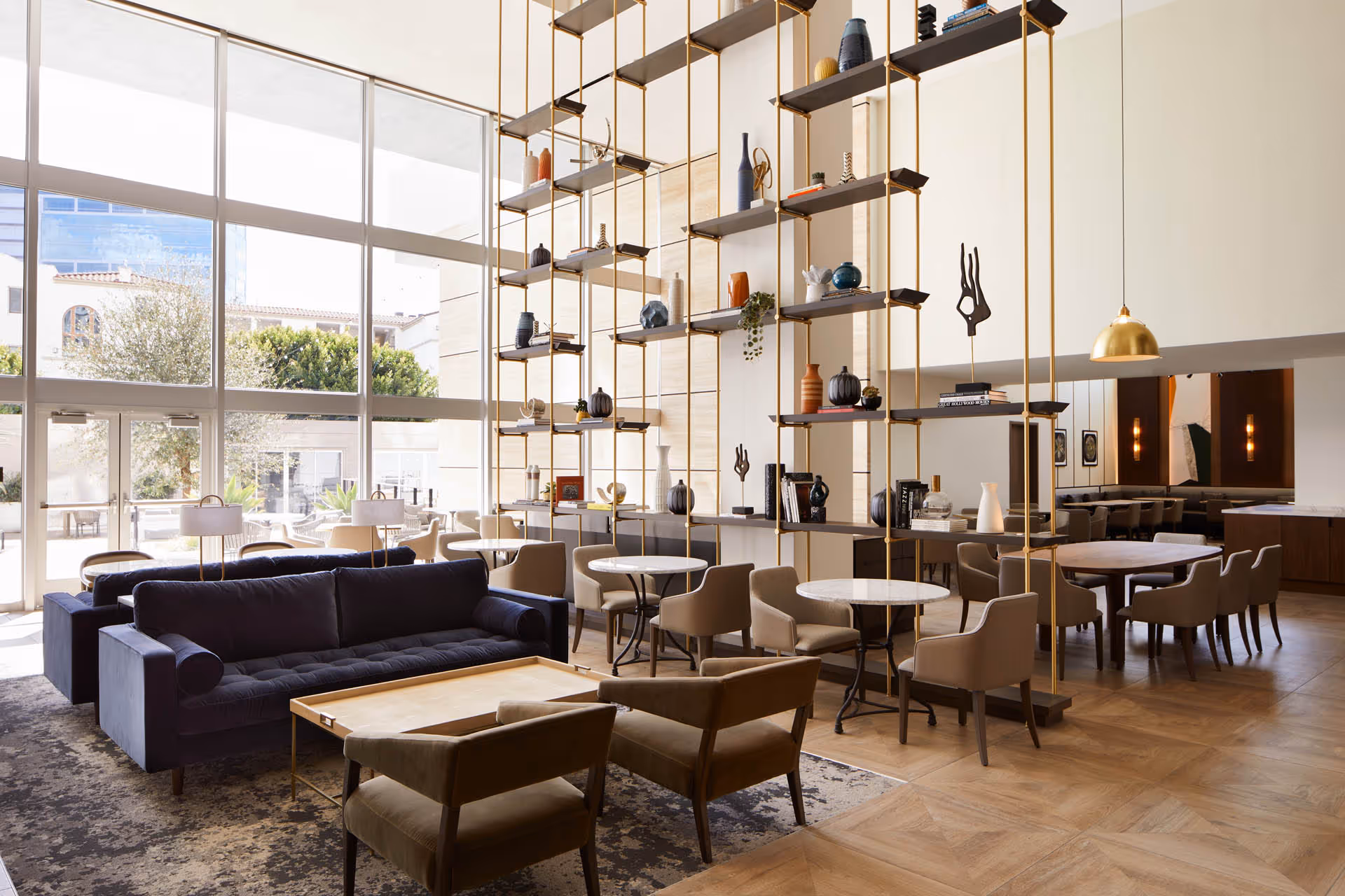 A spacious and modern living room area with large floor-to-ceiling windows letting in natural light. The room features dark blue sofas, beige armchairs, small round marble tables, and a decorative shelving unit with various vases and sculptures. The floor is wooden with a patterned rug under the seating area. In the background, there is a dining area with tables and chairs, and warm lighting fixtures.
