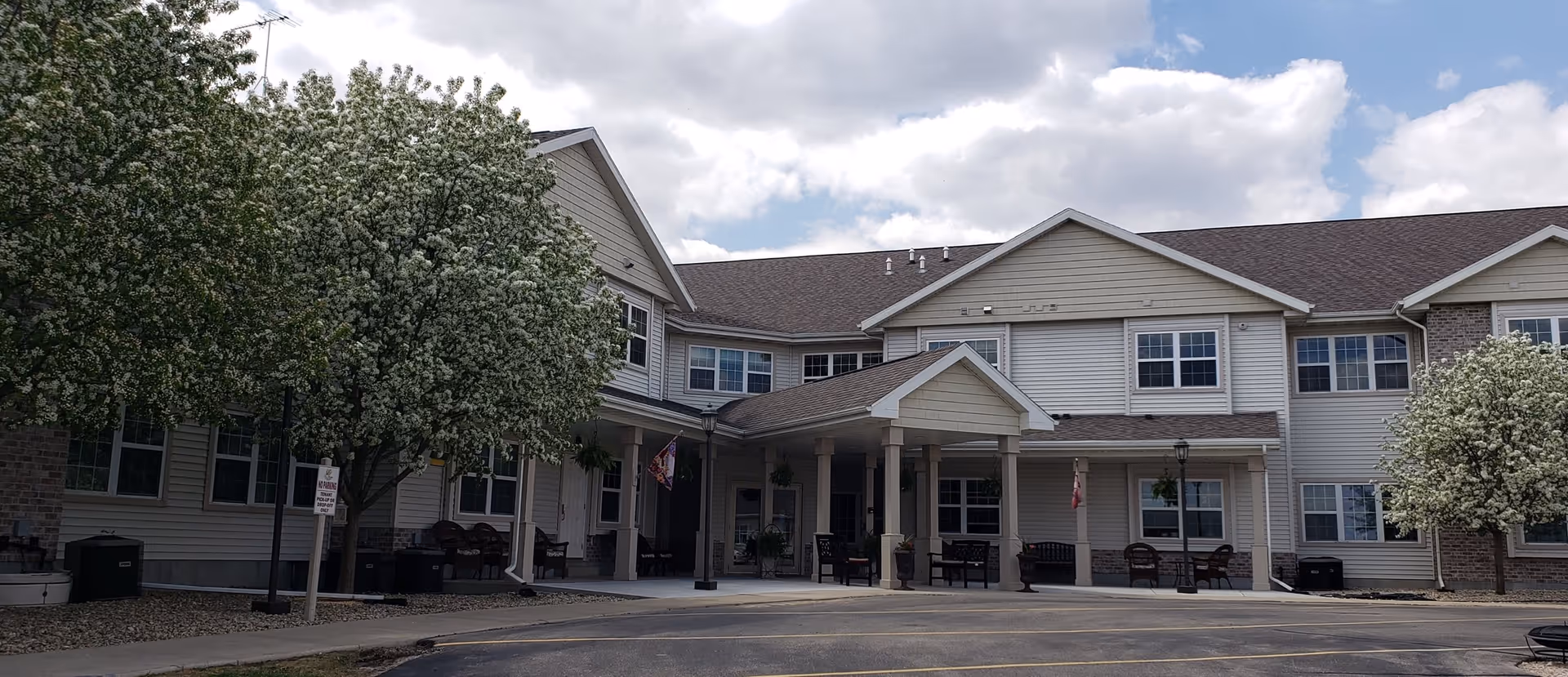 Exterior view of a two-story senior living facility building with beige siding and brick accents. The entrance features a covered porch with columns and several benches. There are blooming trees with white flowers on either side of the entrance, and a paved driveway in front under a partly cloudy sky.