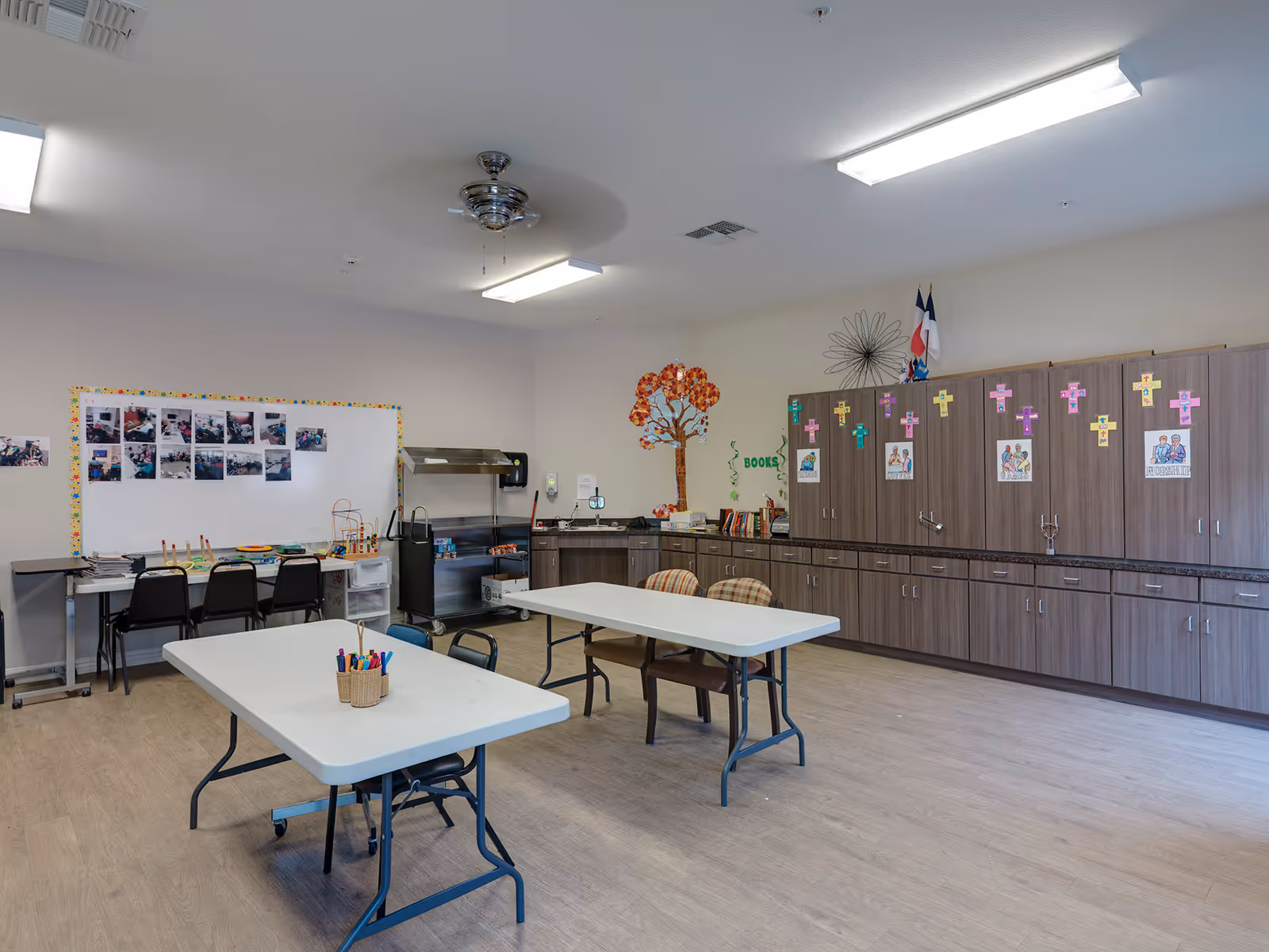 Spacious interior activity room with folding tables and chairs, storage cabinets, a bulletin board and craft supplies.